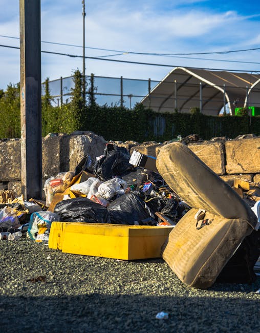 A person wearing white sneakers, navy shorts, and a blue shirt is outdoors on a rocky terrain, using a litter picker to collect small pieces of paper or debris from among large, uneven stones with rou
