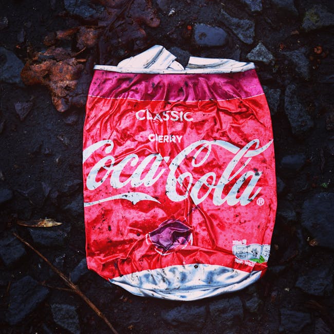 A crumpled, flattened Coca-Cola Classic Cherry drink can with a bright red and white design, including the Coca-Cola logo and cherry imagery, lies on a dark, uneven surface composed of volcanic rocks